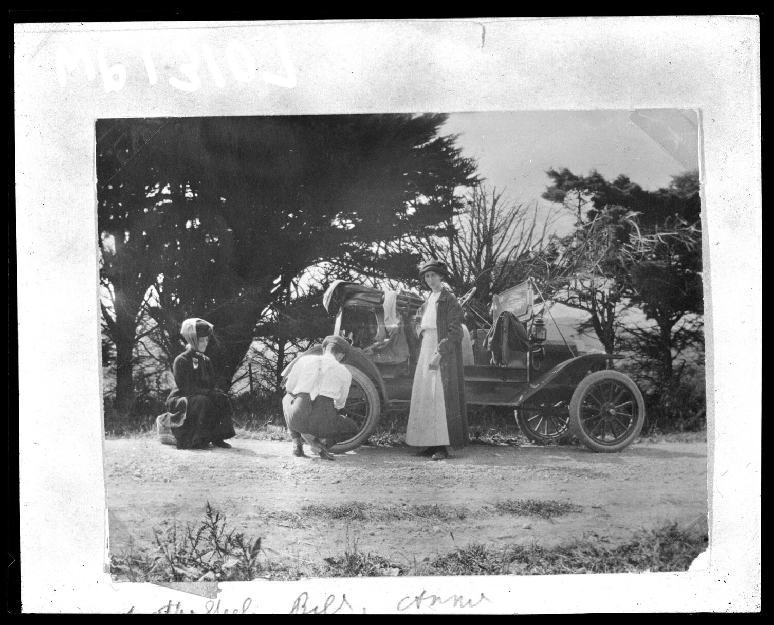 Two women wait while a man repairs the wheel on a motor vehicle