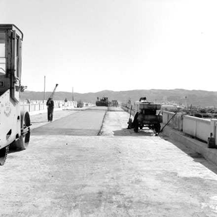 c. Construction of new bridge, workmen, machinery, motor vehicles. Road is being resurfaced