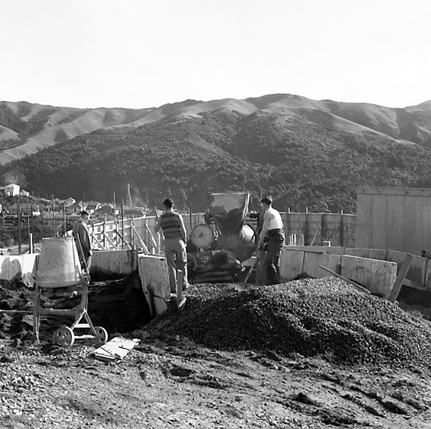 e. View of construction site, workmen are pouring concrete into framing