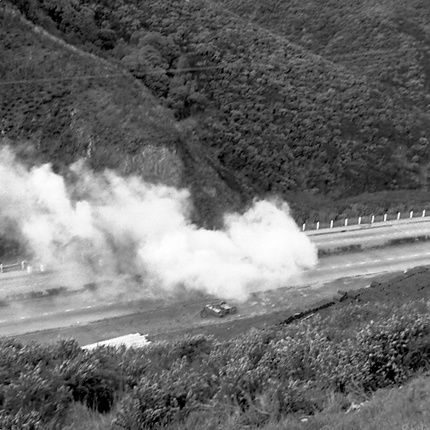 a. Possibly explosives being used to demolish a portion of the hillside. Workmen are preparing site. Elevated view of site with large cloud of dust