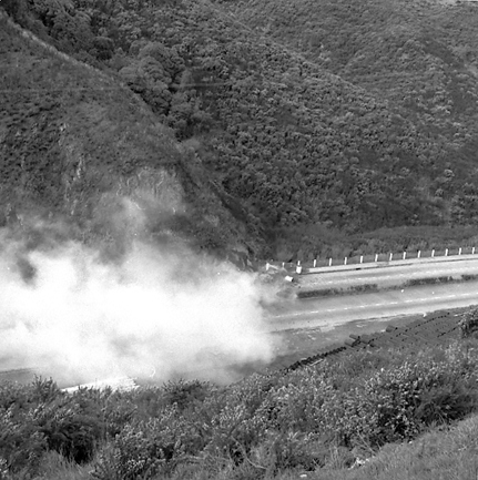 b. Possibly explosives being used to demolish a portion of the hillside. Workmen are preparing site. Elevated view of site with large cloud of dust