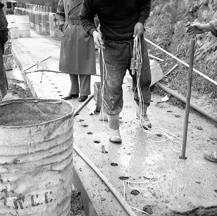 c. Possibly explosives being used to demolish a portion of the hillside. Workmen are preparing site. Elevated view of site with large cloud of dust