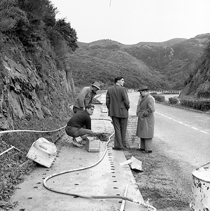 d. Possibly explosives being used to demolish a portion of the hillside. Workmen are preparing site. Elevated view of site with large cloud of dust