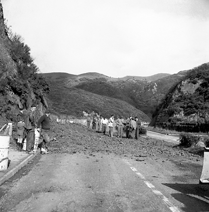 e. Possibly explosives being used to demolish a portion of the hillside. Workmen are preparing site. Elevated view of site with large cloud of dust