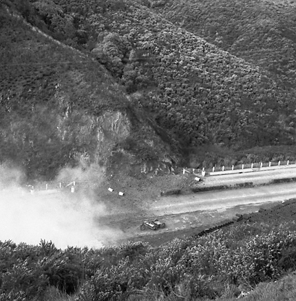 f. Possibly explosives being used to demolish a portion of the hillside. Workmen are preparing site. Elevated view of site with large cloud of dust