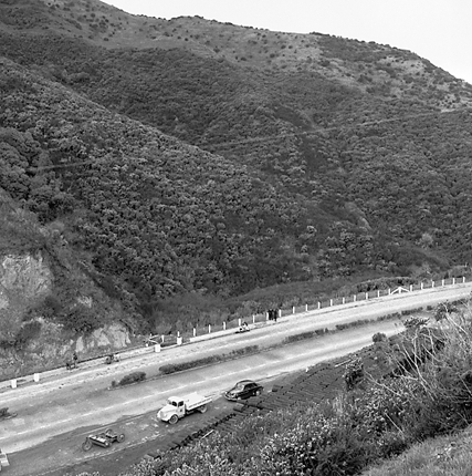g. Possibly explosives being used to demolish a portion of the hillside. Workmen are preparing site. Elevated view of site with large cloud of dust