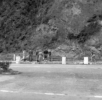 i. Possibly explosives being used to demolish a portion of the hillside. Workmen are preparing site. Elevated view of site with large cloud of dust
