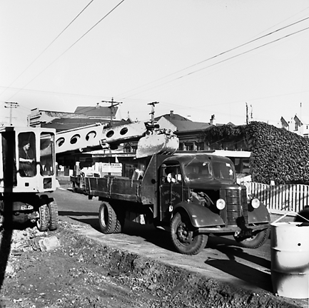 a.Workmen using a front end grader to remove road surface, trucks. Retail buildings in the background