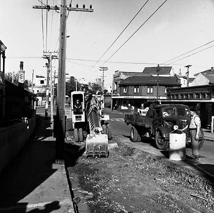 b.Workmen using a front end grader to remove road surface, trucks. Retail buildings in the background