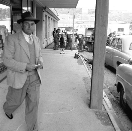 a. Workmen are repairing the footpath outside Willams and Adams Motor Showroom on the corner of Taranaki Street. A girl has stopped to watch the work. Clothing and dress