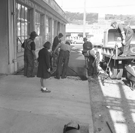 b. Workmen are repairing the footpath outside Willams and Adams Motor Showroom on the corner of Taranaki Street. A girl has stopped to watch the work. Clothing and dress