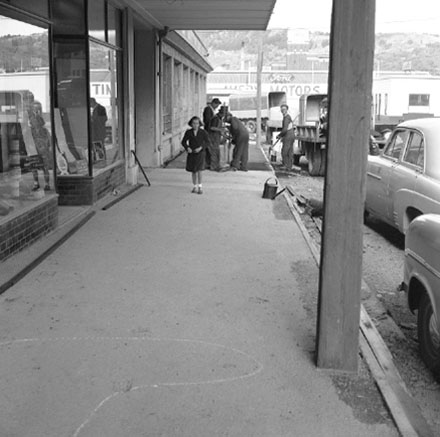 c. Workmen are repairing the footpath outside Willams and Adams Motor Showroom on the corner of Taranaki Street. A girl has stopped to watch the work. Clothing and dress