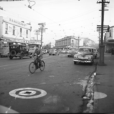 d. John Street and Riddiford Street intersection, pedestrians, tramcars, motor vehicles, retail buildings
