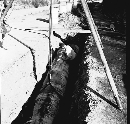 a. Large concrete pipe being laid in a trench. Workmen are using a digger to dig the trench and load soil into the back of a truck . Contractor is C R Quinn