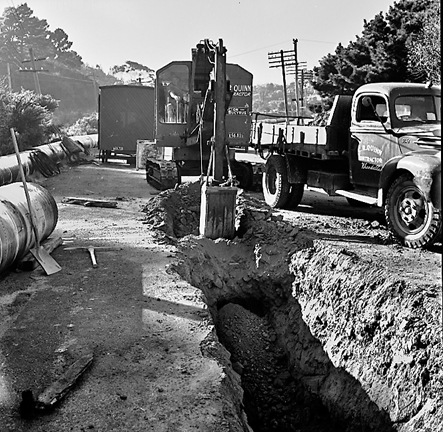b. Large concrete pipe being laid in a trench. Workmen are using a digger to dig the trench and load soil into the back of a truck . Contractor is C R Quinn