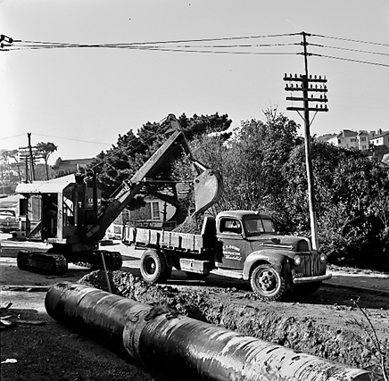 c. Large concrete pipe being laid in a trench. Workmen are using a digger to dig the trench and load soil into the back of a truck . Contractor is C R Quinn