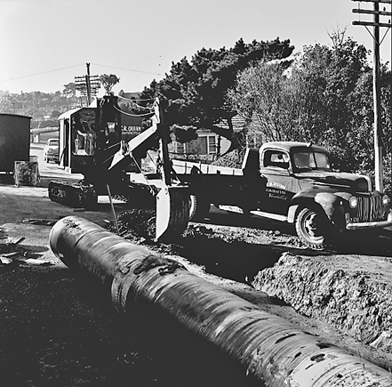 d. Large concrete pipe being laid in a trench. Workmen are using a digger to dig the trench and load soil into the back of a truck . Contractor is C R Quinn
