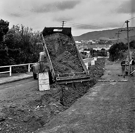 a. Large concrete pipe being laid in a trench. Workmen are using a digger to dig the trench and transfer the pipe from the back of a truck. Contractor is C R Quinn