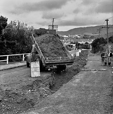 b. Large concrete pipe being laid in a trench. Workmen are using a digger to dig the trench and transfer the pipe from the back of a truck. Contractor is C R Quinn
