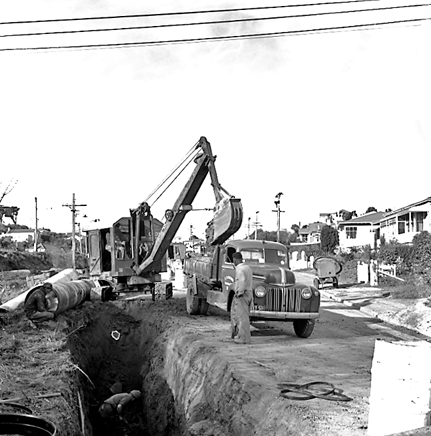 d. Large concrete pipe being laid in a trench. Workmen are using a digger to dig the trench and transfer the pipe from the back of a truck. Contractor is C R Quinn