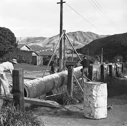 e. Large concrete pipe being laid in a trench. Workmen are using a digger to dig the trench and transfer the pipe from the back of a truck. Contractor is C R Quinn