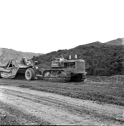d. views of construction work in progress, workmen, equipment, graders, front end loaders, trucks, dwellings in the background