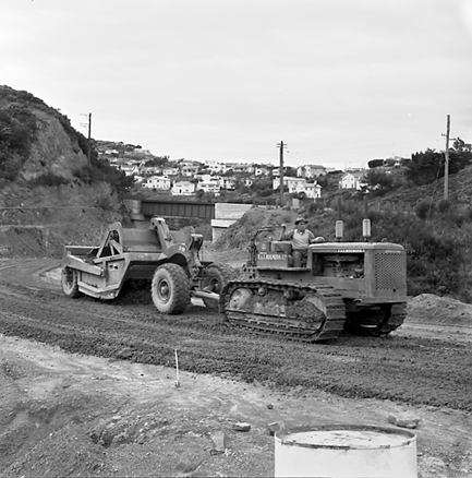i. views of construction work in progress, workmen, equipment, graders, front end loaders, trucks, dwellings in the background