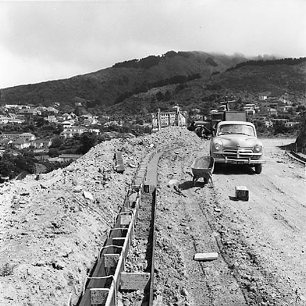 a. Road and kerb construction, motor vehicle in foreground. Workmen having break in background