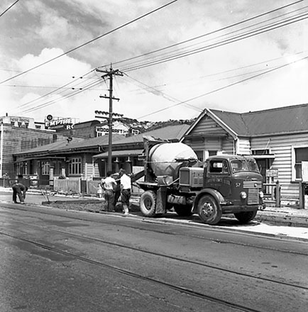 a. Workmen are pouring concrete from ' Certified Concrete Ltd ' truck to seal a piece of road in front of 8 and 10 Adelaide Road, a single storey wooden dwelling and Saint Annes Daycare