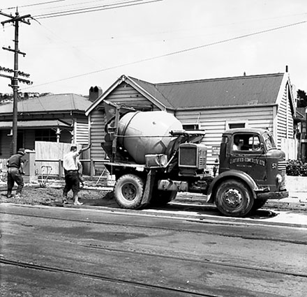 b. Workmen are pouring concrete from ' Certified Concrete Ltd ' truck to seal a piece of road in front of 8 and 10 Adelaide Road, a single storey wooden dwelling and Saint Annes Daycare