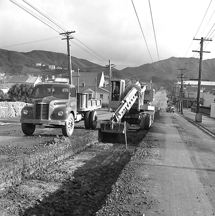 a. Workmen are using a Gradall to dig a trench and load the excess soil on to trucks. Trolley buses passing by