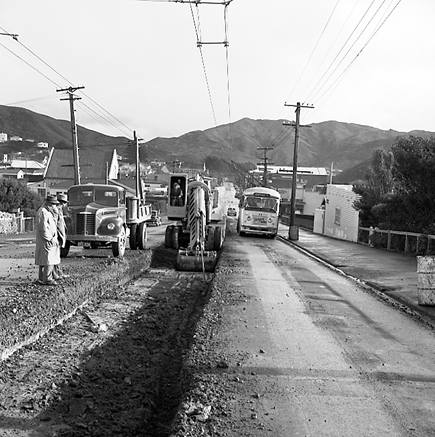 b. Workmen are using a Gradall to dig a trench and load the excess soil on to trucks. Trolley buses passing by