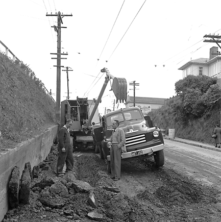 d. Workmen are using a Gradall to dig a trench and load the excess soil on to trucks. Trolley buses passing by