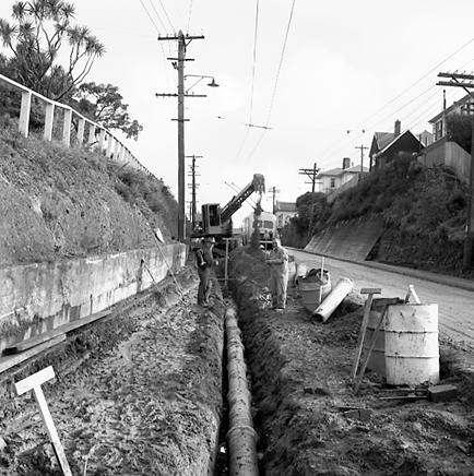 e. Workmen are using a Gradall to dig a trench and load the excess soil on to trucks. Trolley buses passing by