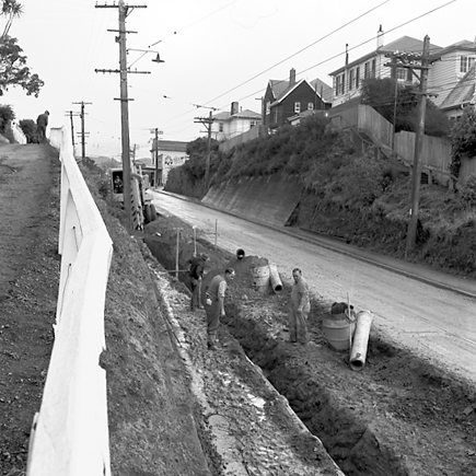 f. Workmen are using a Gradall to dig a trench and load the excess soil on to trucks. Trolley buses passing by