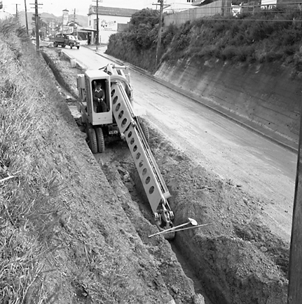 g. Workmen are using a Gradall to dig a trench and load the excess soil on to trucks. Trolley buses passing by