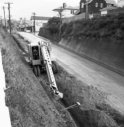 h. Workmen are using a Gradall to dig a trench and load the excess soil on to trucks. Trolley buses passing by