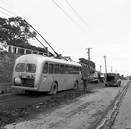 i. Workmen are using a Gradall to dig a trench and load the excess soil on to trucks. Trolley buses passing by