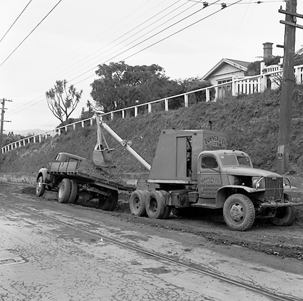 j. Workmen are using a Gradall to dig a trench and load the excess soil on to trucks. Trolley buses passing by