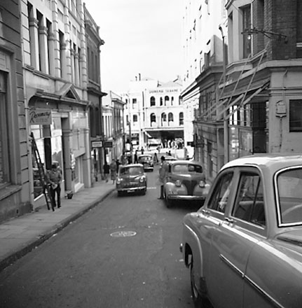 a. Woodward Street, shop fronts, motor vehicles, workman is washing windows on the left. Looking east towards Lambton Quay