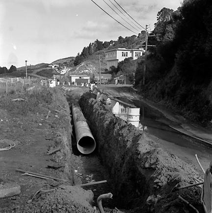 c. Intersection with Travers Street. Road works are in progress and a front end digger is being used to dig a trench. Workmen and machinery, drainage pipes are ready to be laid