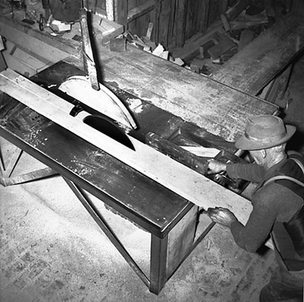 b. Interior of workshop. Workman cutting wood with a table saw