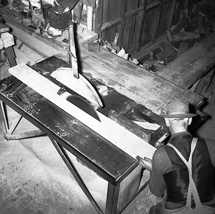 c. Interior of workshop. Workman cutting wood with a table saw