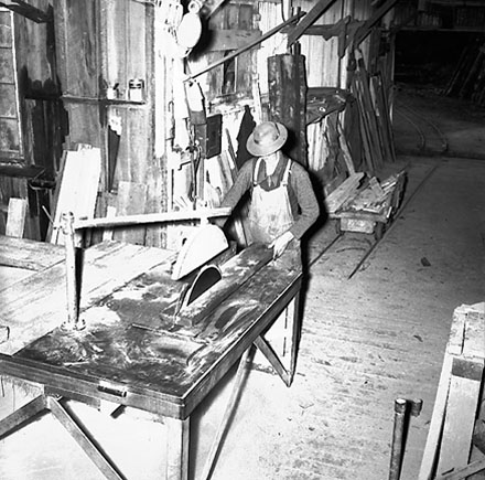 e. Interior of workshop. Workman cutting wood with a table saw