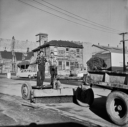 a. Two workmen standing on back of road surfacing equipment. Rundown dwellings in the background