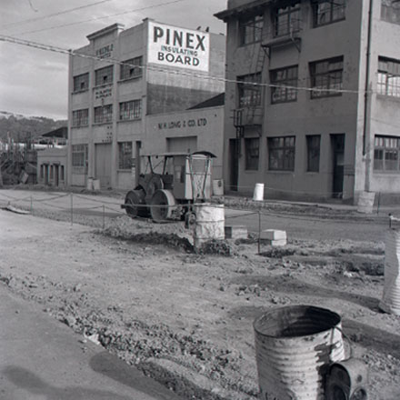 a . Views of road work being carried out by the Drainage Department. Excavation equipment and roller. In the background, W H Long &amp; Co, Foresters Arms