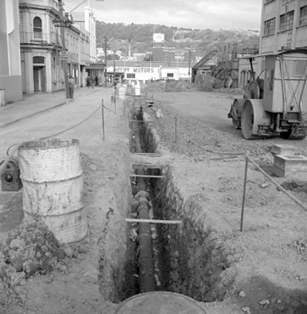 b . Views of road work being carried out by the Drainage Department. Excavation equipment and roller. In the background, W H Long &amp; Co, Foresters Arms