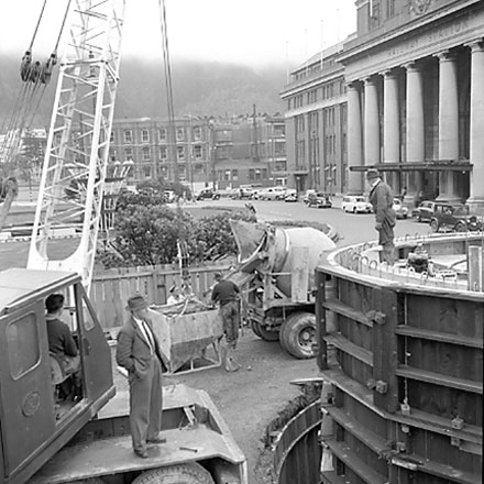 a. views of work in progress, workmen pouring concrete using an overhead crane. Railway Station in background