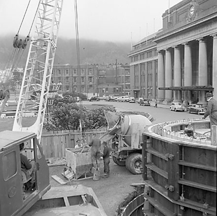 b. views of work in progress, workmen pouring concrete using an overhead crane. Railway Station in background