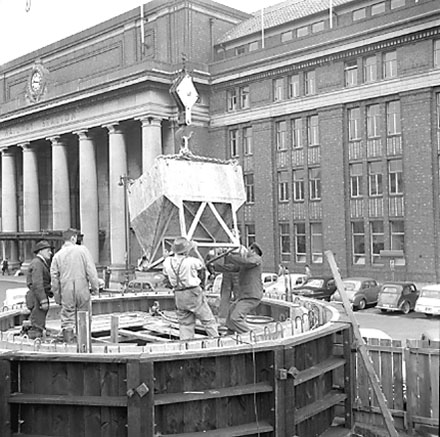 c. views of work in progress, workmen pouring concrete using an overhead crane. Railway Station in background